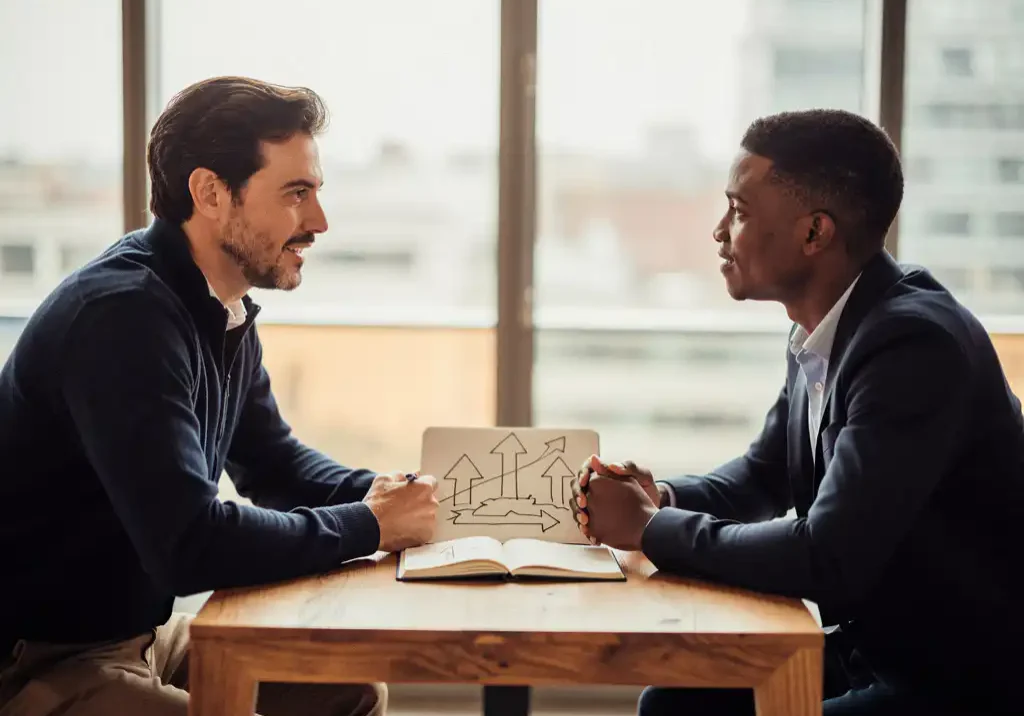 Coach en cliënt in gesprek aan houten tafel met opengeslagen notitieboek met groeidiagram, warm kantoor met natuurlijk daglicht.